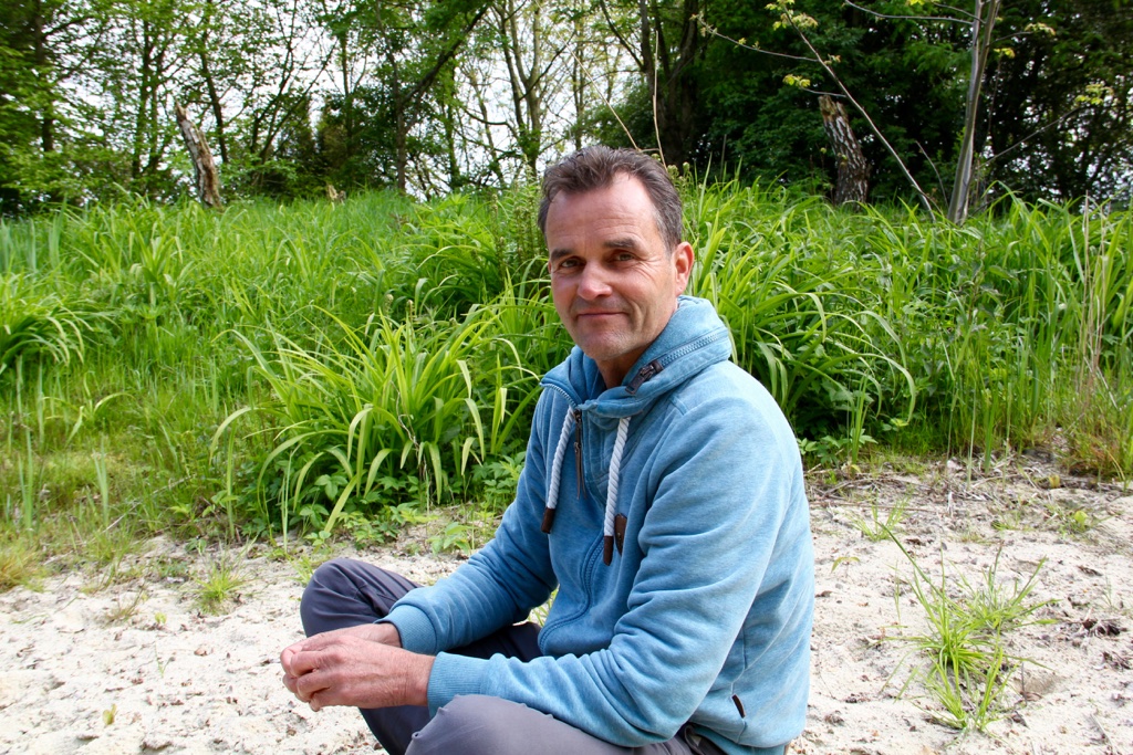 Volker Kranz sitzt am sandigen Strand des kleinen Teichs im Essgarten. Hinter ihm wachsen feuchtigkeitsliebende Gräser und andere Pflanzen.